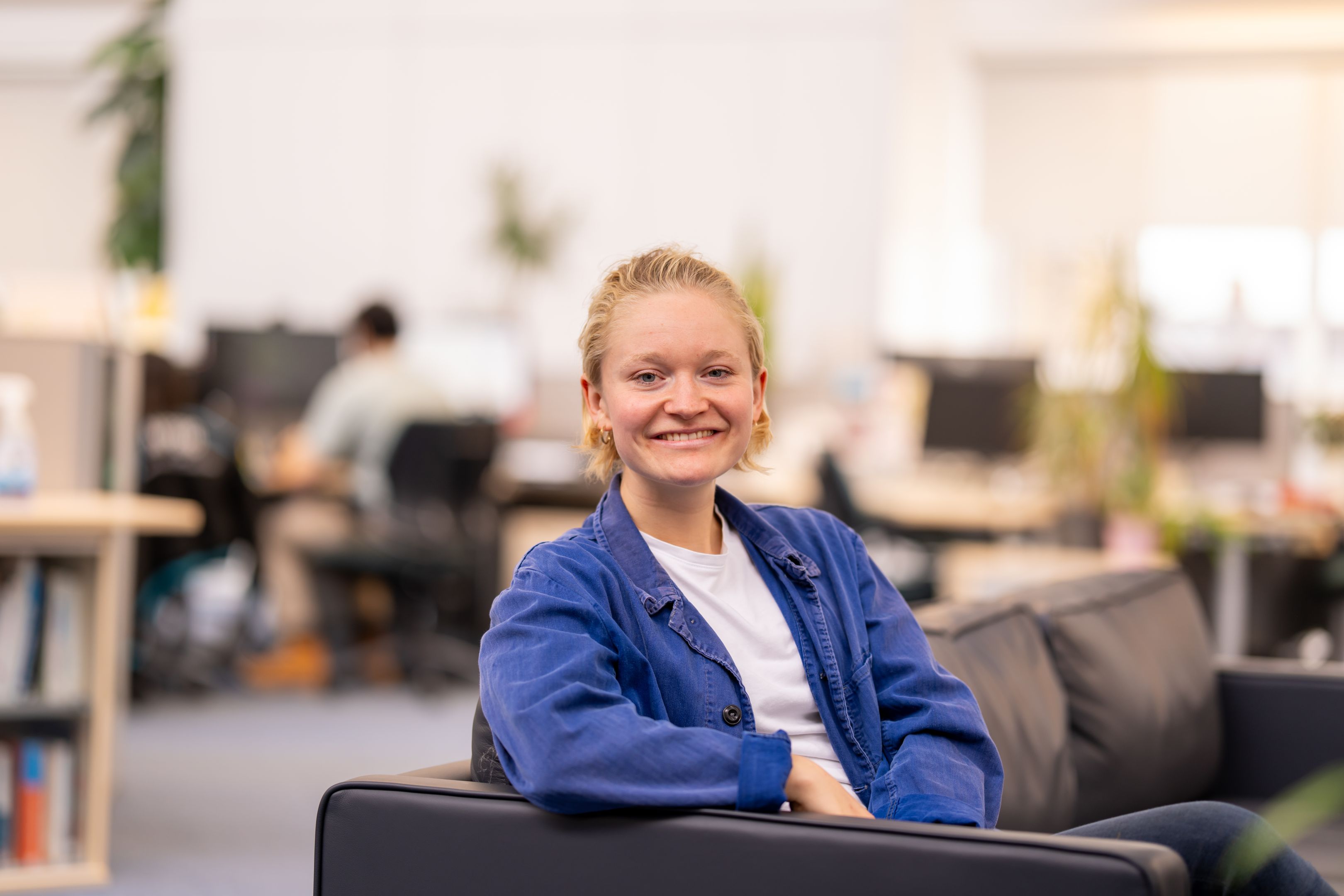 A smiling woman with blond hair and wearing a blue blazer sitting on black sofa. 