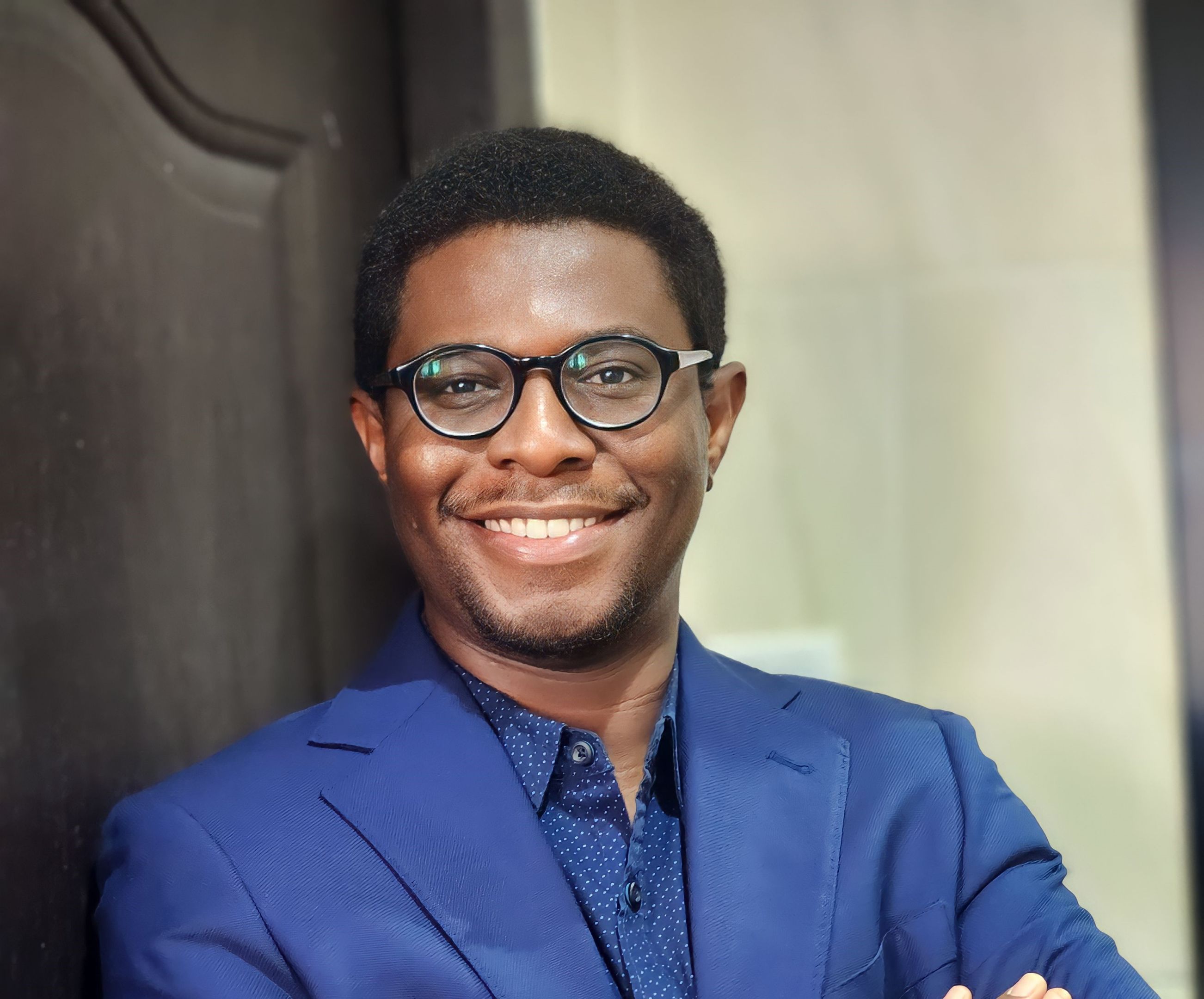 A smiling man leaning against a door. He is wearing, blackrimmed glasses, a blue shirt and suit. 
