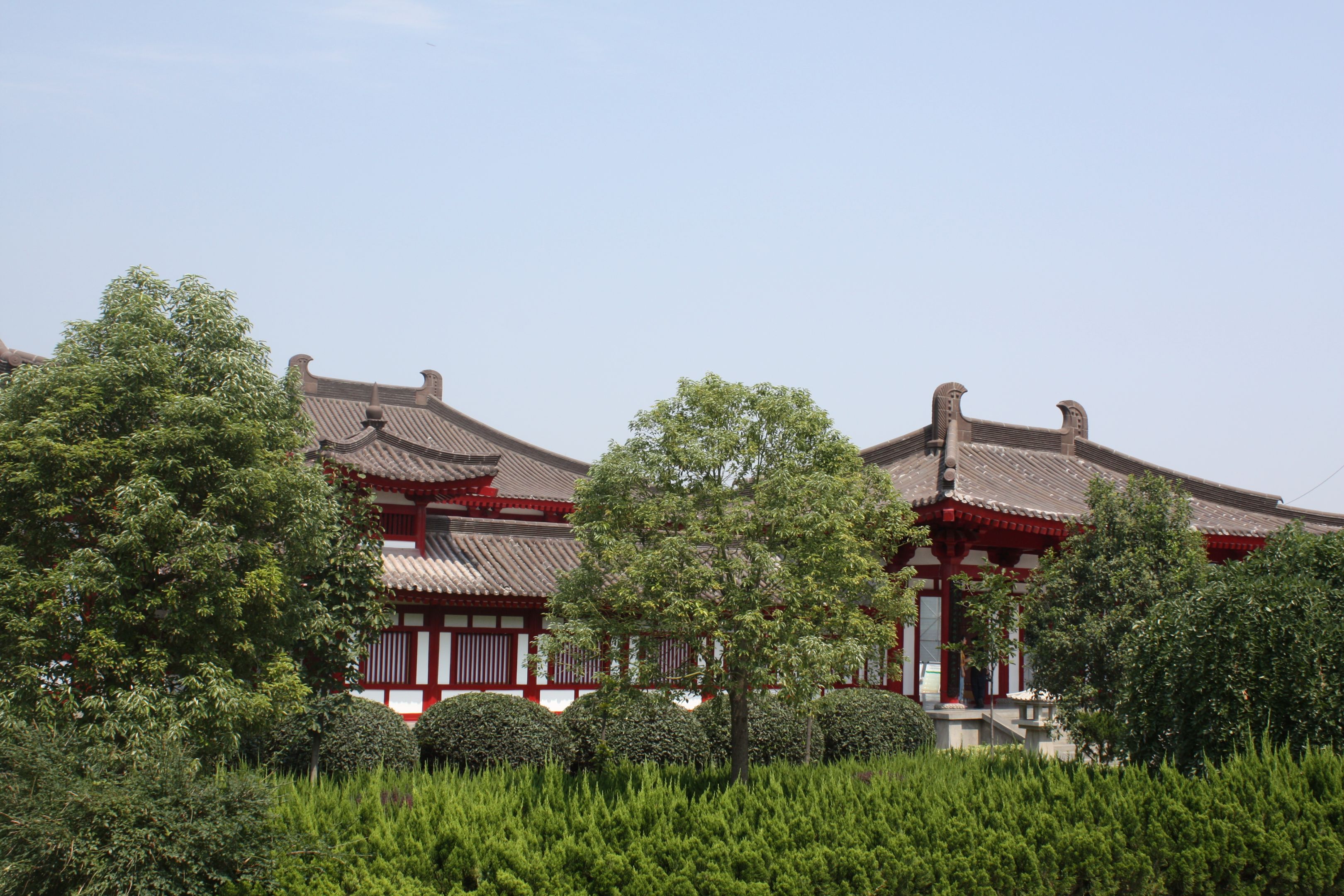 Buildings with grey roof tiles, white wall and red- painted detailing behind a leafy, green trees