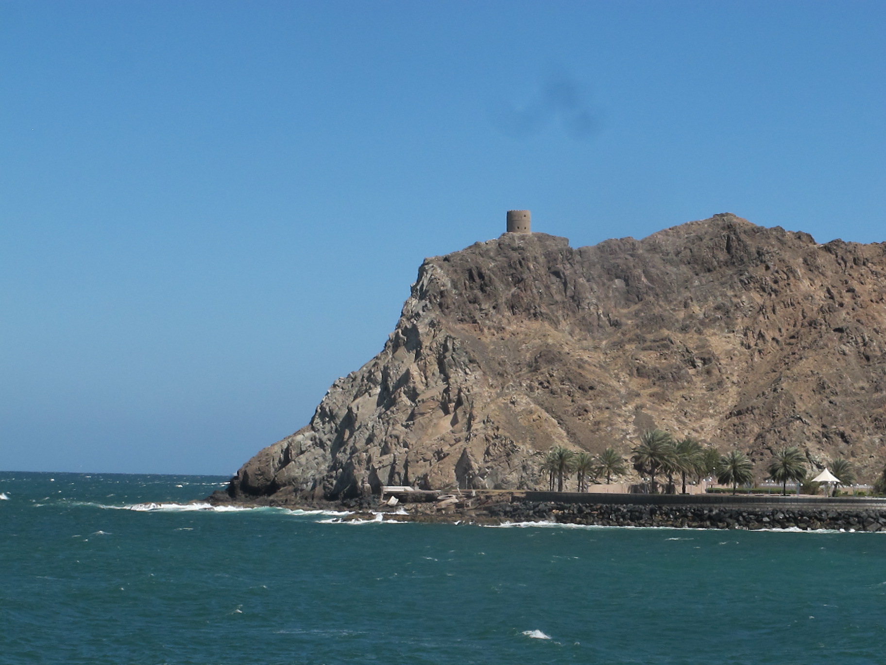 A rocky outcross with small, stone tower on top, jutting out into the a choppy sea, with blue sky in background