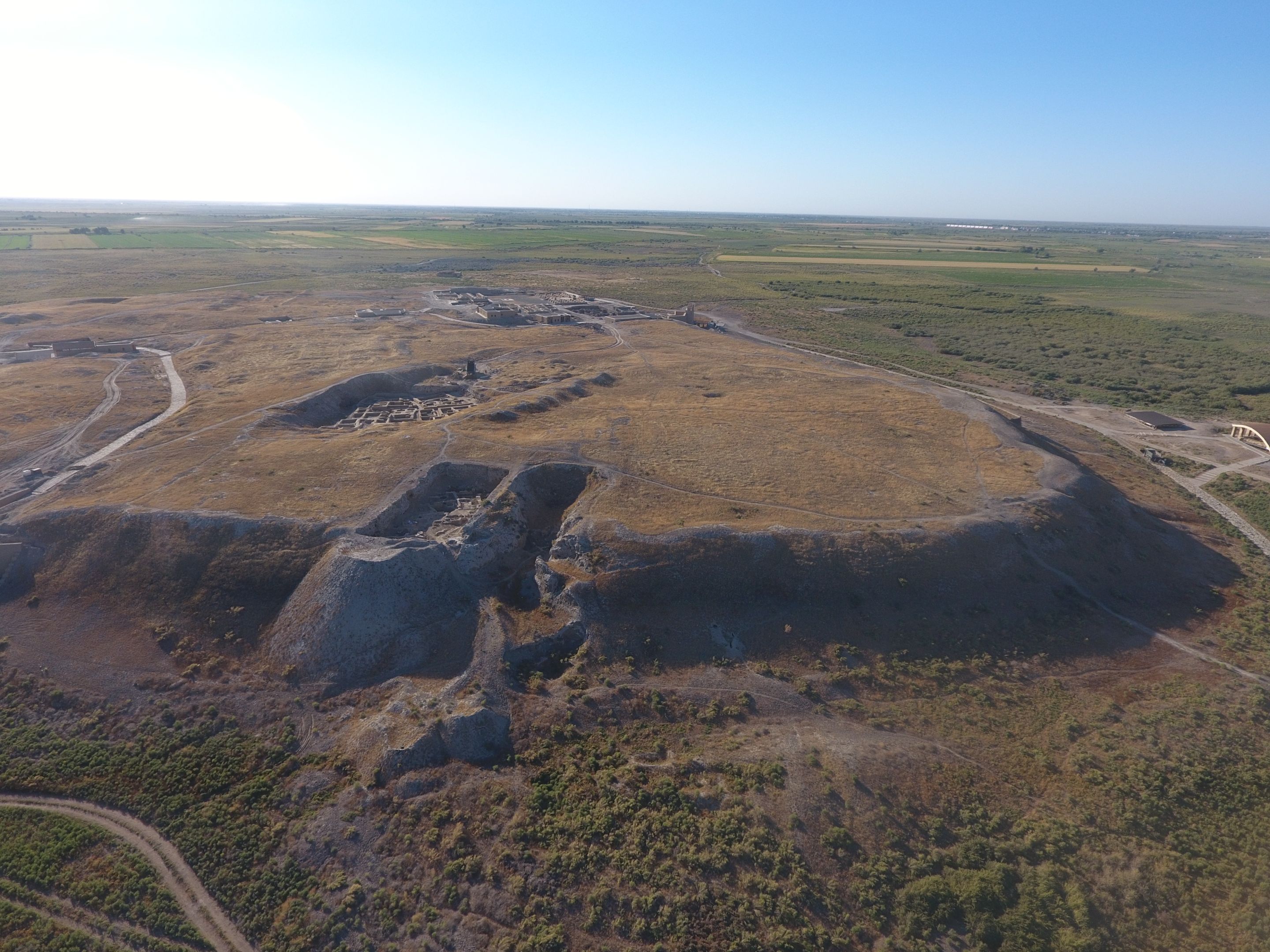 Aerial photo of the archaeological mound of Otrartobe in southern Kazakhstan, with surrounding brownish green vegetation and blue sku at the top of the photo. Large archaeological trenches are visible in darker patches on the mound