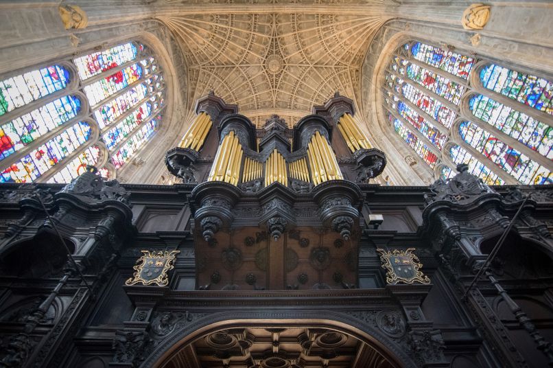 A large organ with gold pipes and dark wood surrounded by stained glass windows with elaborate stone detail