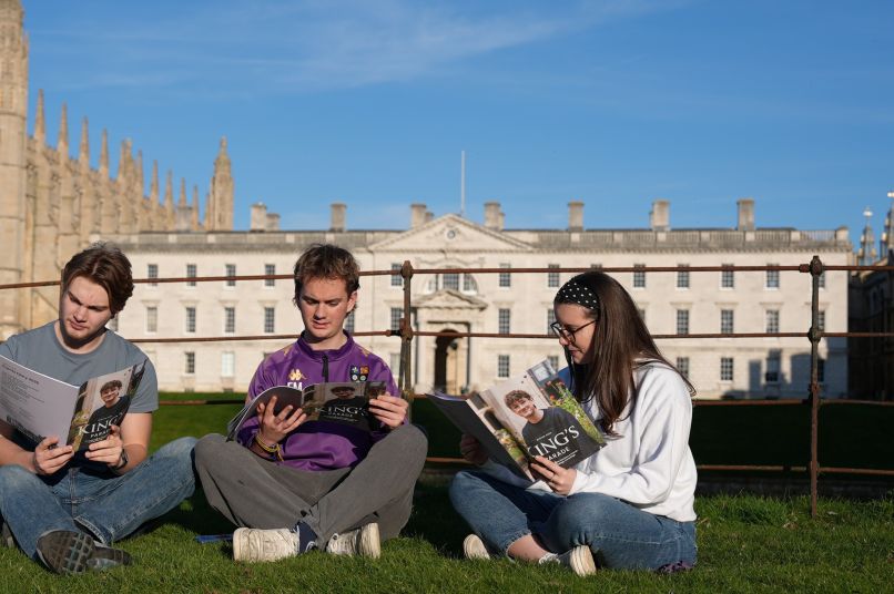 Students reading a magazine by the river with College building behind them