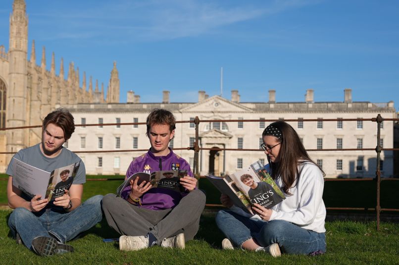 Three students sitting on a grassy river bank reading King's Parade magazine with the Chapel and the Gibbs building in the background