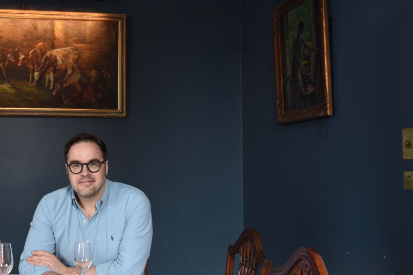 A man with dark hair and glasses sits at a table in a room with dark blue walls and an oil painting behind him
