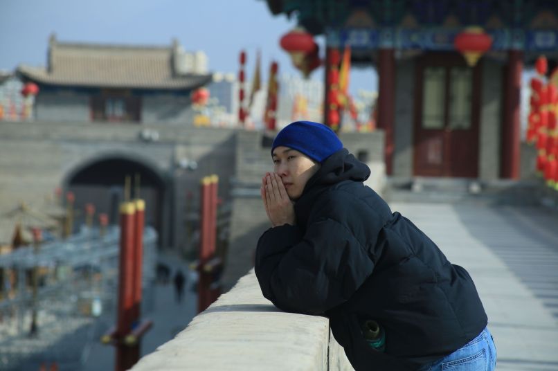 A man in a black winter coat leaning on a low wall with buildings in chinese style in the background.