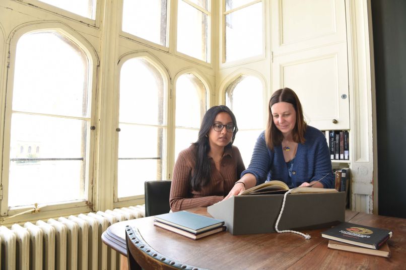 Two women sitting in front of large windows looking at a book together