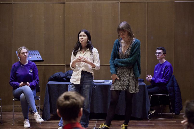 Two students stood on stage speaking with two other students sat in the background with purple t-shirts