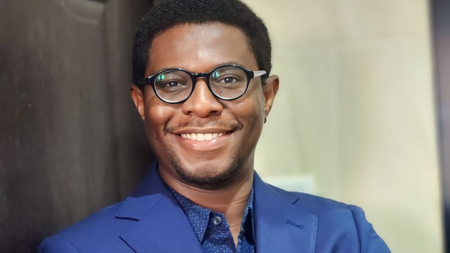 A smiling man leaning against a door. He is wearing, blackrimmed glasses, a blue shirt and suit. 