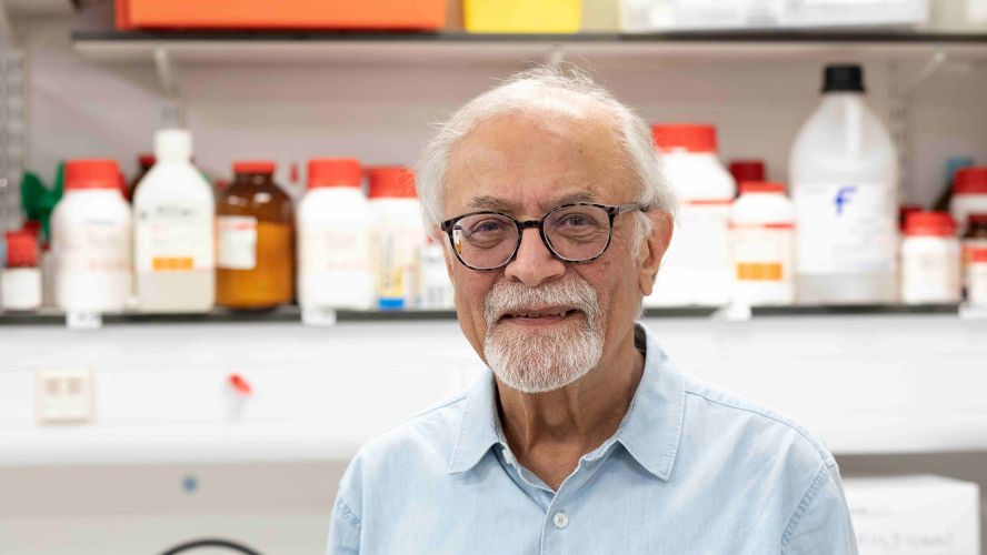 A smiling man with short white hair, a beard and glasses, wearing a pale blue shirt in a lab with bottles on a shelf behind him.