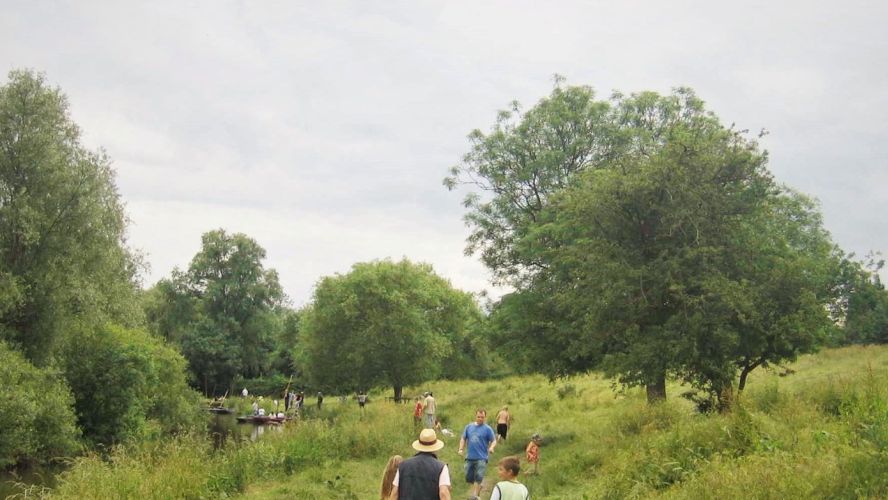 A meadow with a river to the left, people punting and other walking on the bank