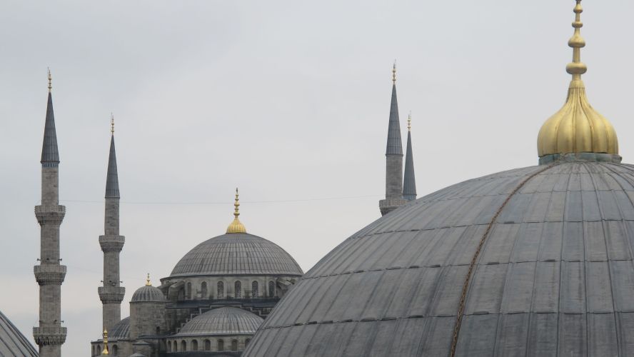 Grey domes and minarets in the ottoman style, one with golden finial, against a grey sky