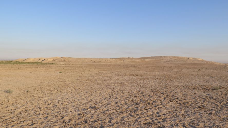 Arid desert landscape with archaeological mound in the distance, blue sky above