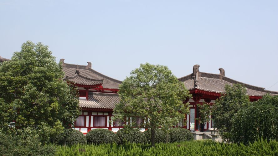 Buildings with grey roof tiles, white wall and red- painted detailing behind a leafy, green trees