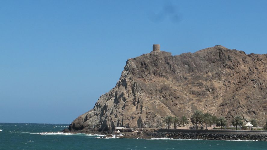A rocky outcross with small, stone tower on top, jutting out into the a choppy sea, with blue sky in background