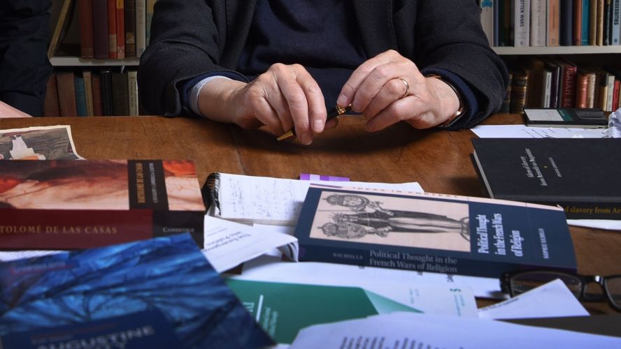 a table covered in books with a pair of hands holding a pen. 