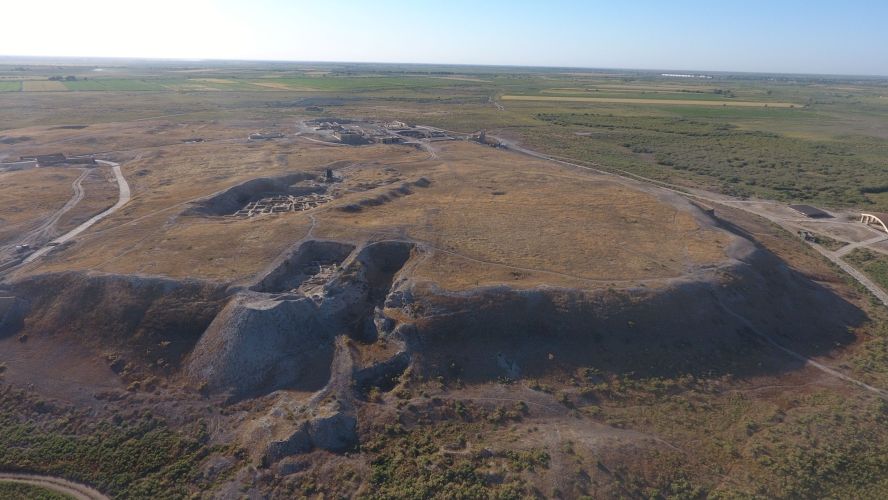 Aerial photo of the archaeological mound of Otrartobe in southern Kazakhstan, with surrounding brownish green vegetation and blue sku at the top of the photo. Large archaeological trenches are visible in darker patches on the mound