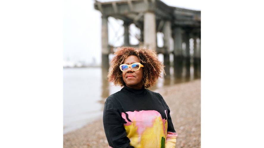 A woman curly hair and yellow glasses stands beside a pier 
