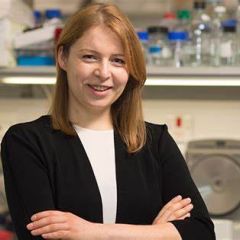 A woman with shoulder length light hair, wearing a black blazer, standing with arms crossed in a lab.
