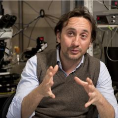 A man with a brown jumper and blue shirt, talking animatedly, with laboratory instruments in the background.