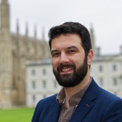 A bearded man, smiling, wearing a blue blazer, in front of the King's College Chapel and lawn.