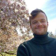 A smiling man with blond hair and beard with a blooming tree in the background.
