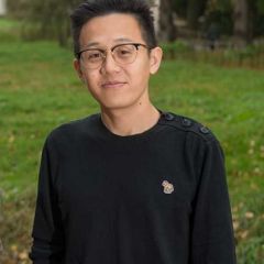A smiling man with dark hair and glasses, standing in a green meadow.