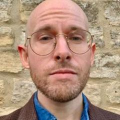A man with glasses with a short beard in front of a stone wall.