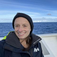 A smiling woman wearing a waterproof dark jacket and black knitted hat on a boat with the sea and blue sky in the background.
