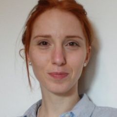 A smiling woman with red hair, wearing a blue collared shirt in front of a white background. 