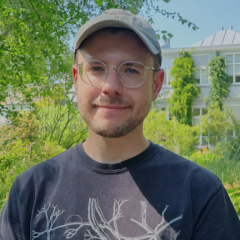 A picture of a smiling man, wearing glasses and a baseball cap, with trees and an ivy covered building in the background.
