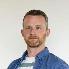 A man with short blond hair and beard, wearing a blue collared shirt with a stripey t-shirt underneath standing in front of a beige background.