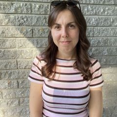 A smiling woman with long hair, wearing a black ans white stripey t-shirt in front of a grey wall. 