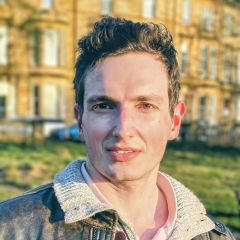 A man with a dark jacket over a lighter, knitted jumper, standing in the sunshine outside a city building. 