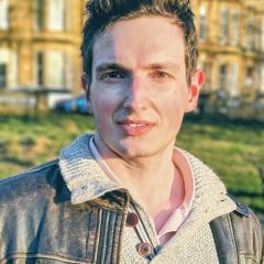 A man with a dark jacket over a lighter, knitted jumper, standing in the sunshine outside a city building.