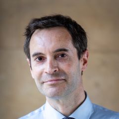 Jason Sharman, a smiling man with short dark hair. He is wearing a blue shirt and a blue pattered tie.