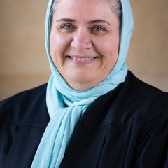 Mezna Qato, a smiling woman with grey hair wearing a light blue headscarf and a black robe. 