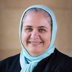 Mezna Qato, a smiling woman with grey hair wearing a light blue headscarf and a black robe. 