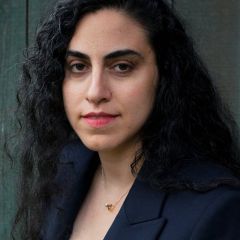 A woman with long, dark hair and wearing a blue blazer in front of a dark background.