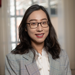 A smiling woman with shoulder length dark hair, wearing glasses and a grey blazer over a white shirt.