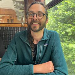 A smiling bearded man with glasses, sitting in a train. 