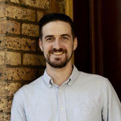 smiling man with dark hair leaning against a brick wall. 