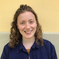 A smiling woman with curly, shoulder length dark blonde hair, wearing a dark blue collared shirt open at the neck, in front of a yellow and beige background.