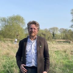smiling man standing in a field wearing a brown suit jacket and white shirt