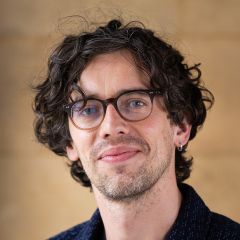 Phil Knox, a smiling man with thick dark curly hair. He is wearing glasses, a small silver earring and a casual blue shirt. 