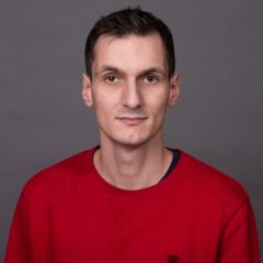 A man with short, dark hair wearing a red jumper in front of a grey background. 