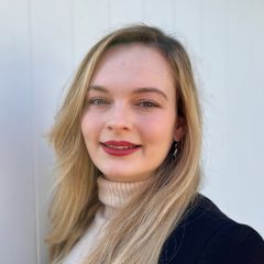 Sarah Paris, a smiling woman with long hair, wearing a white turtleneck jumper and a black jacket. 