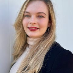 Sarah Paris, a smiling woman with long hair, wearing a white turtleneck jumper and a black jacket. 