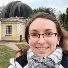 A smiling woman with glasses and long hair, wearing a scarf and jacket in front of an observatory. 