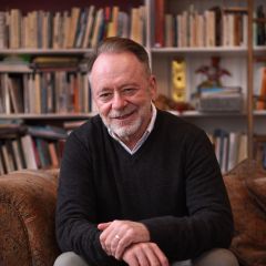 a man sits on a sofa with a full bookcase behind him smiling at the camera